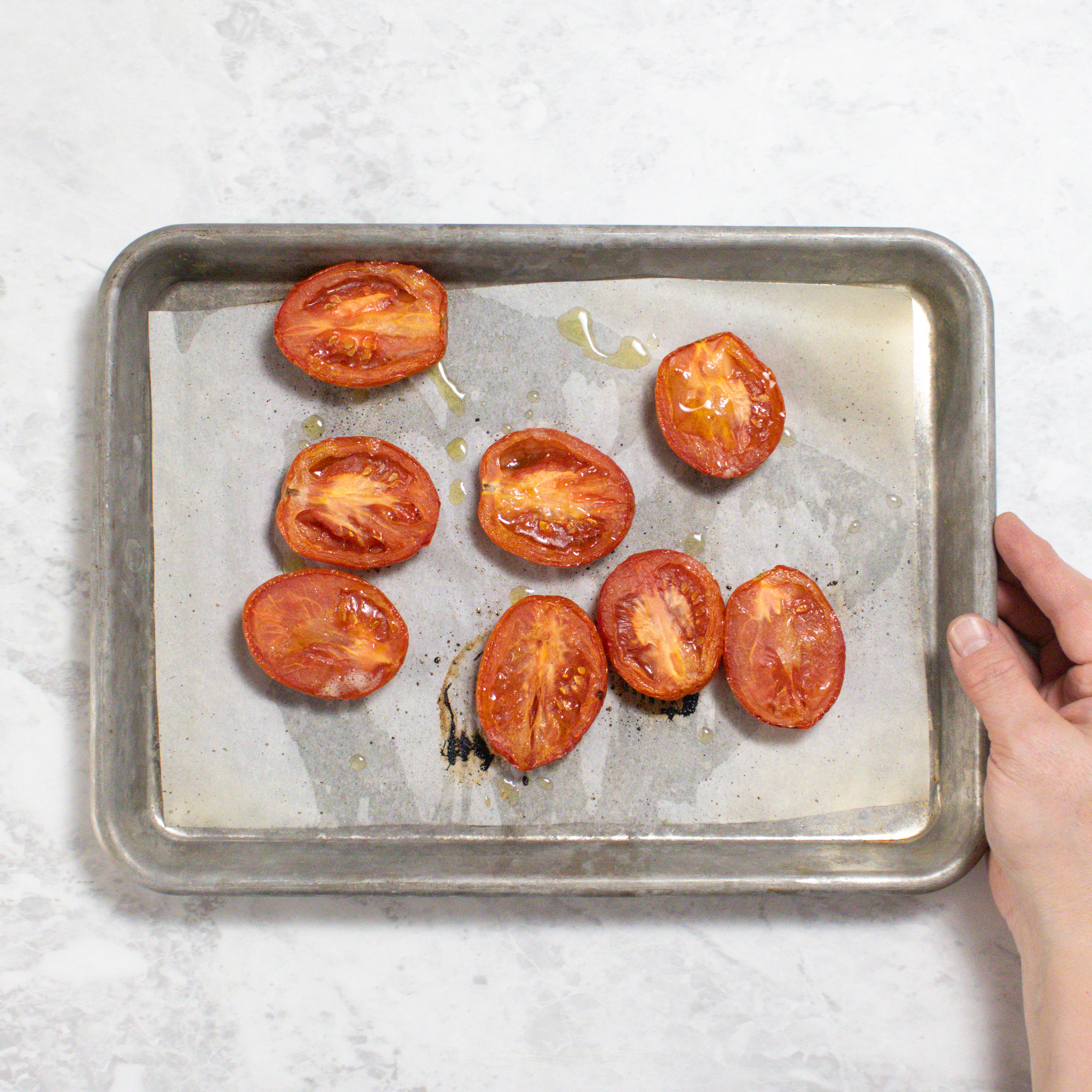 Hand holding a silver baking tray on my white marble kitchen counter. The tomatoes have been roasted.