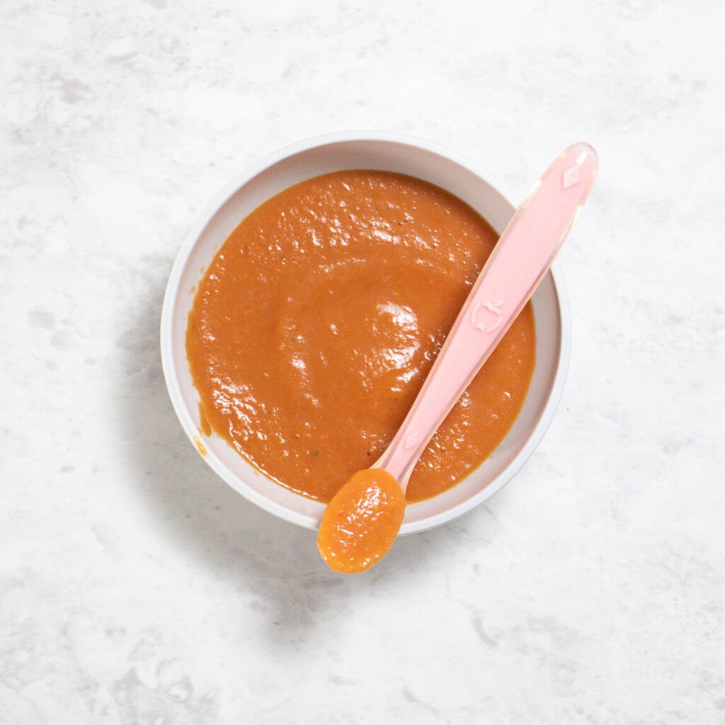 Gray baby bowl with pureed tomatoes and basil sitting on a marble counter with a pink baby spoon.