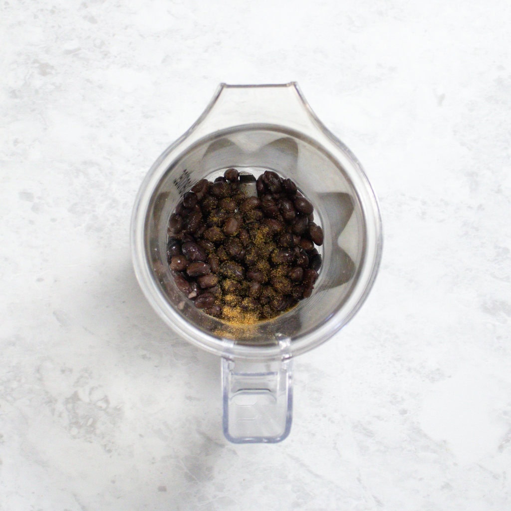 small clear blender full of black beans and pinch of cumin sitting on my white marble counter.