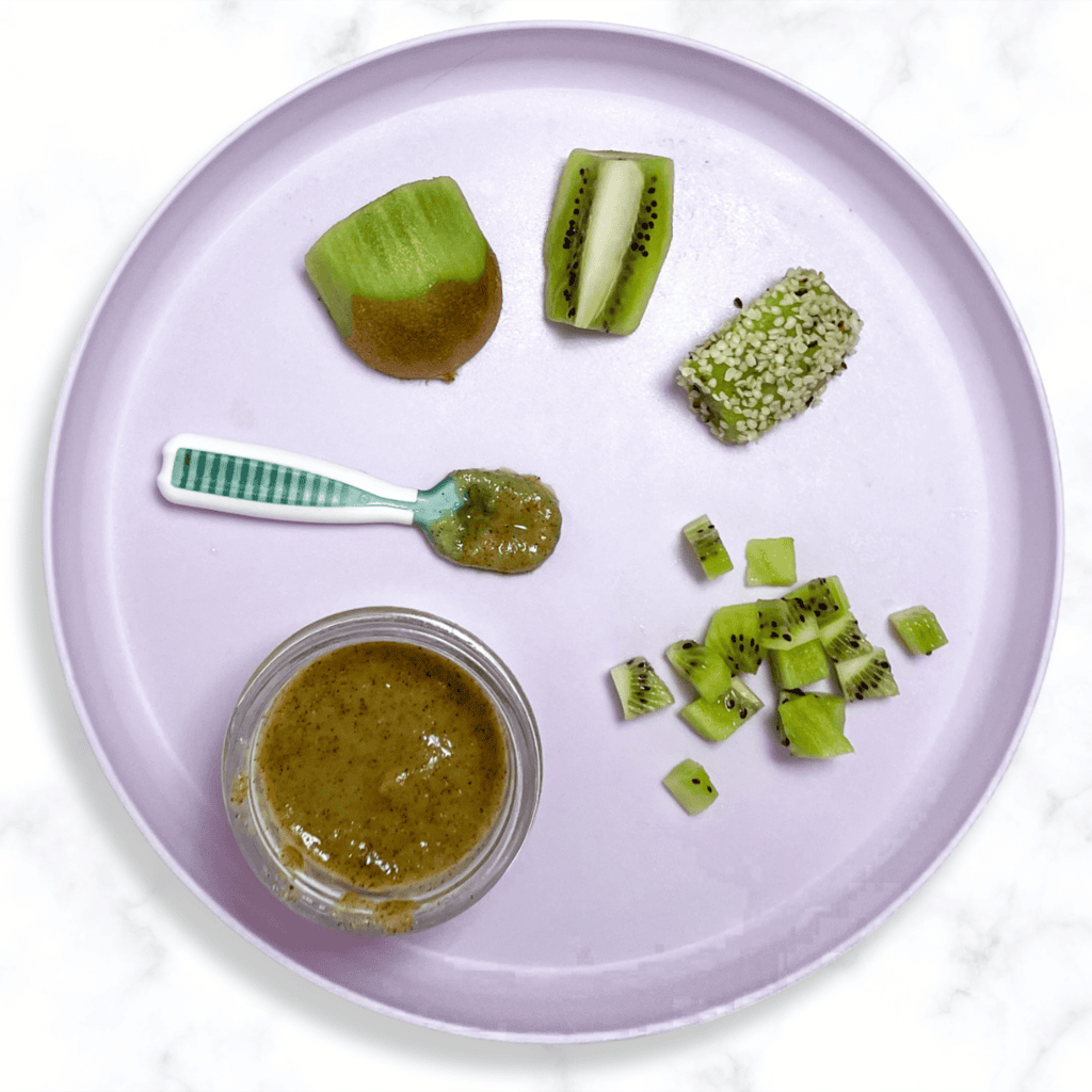 Purple baby plate on my white kitchen counter, with kiwi cut and pureed in different ways showing how you can serve it at different ages and stages. 