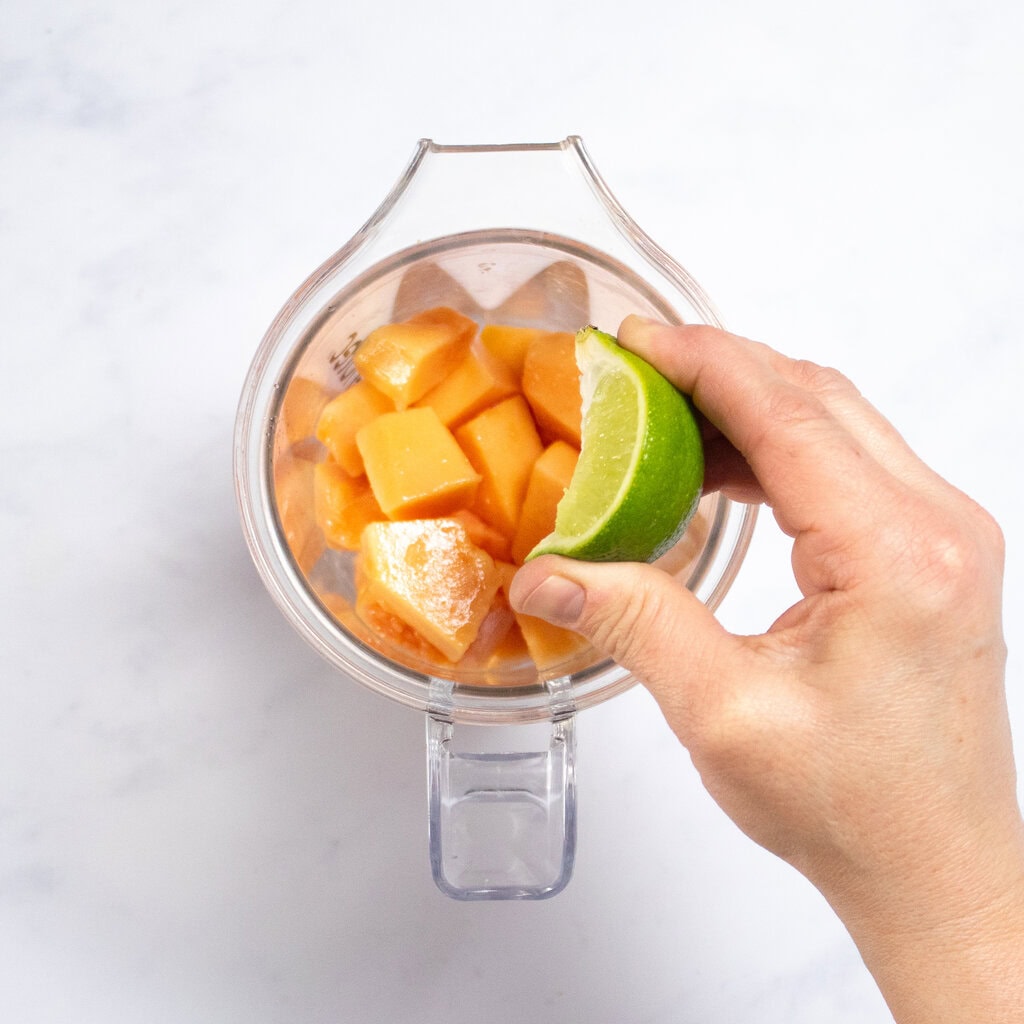 Clear blender on my counter with chunks of papaya and my hand squeezing lime inside.