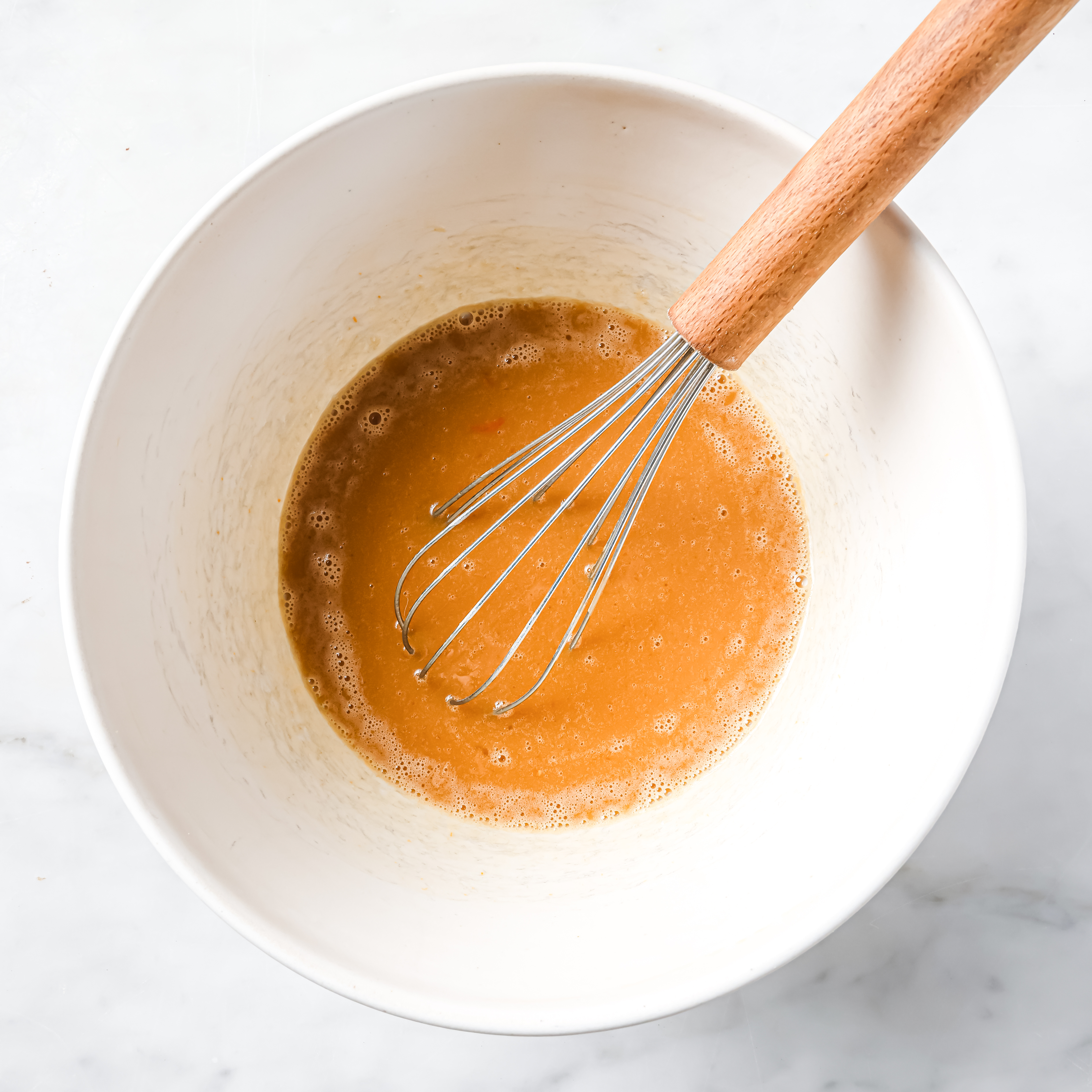 White mixing bowl on kitchen counter with wet ingredients and whisk resting inside.