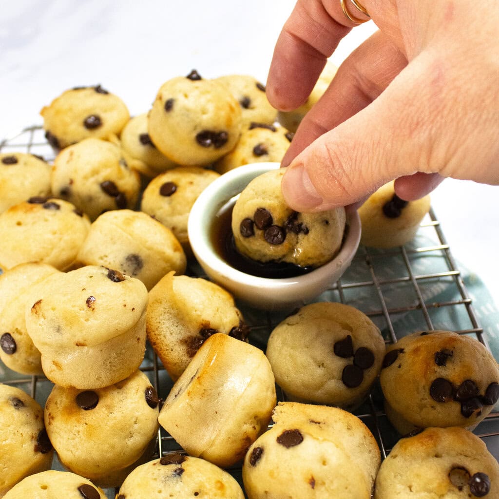My hand dunking a mini pancake bite into a small bowl of maple syrup with other bites resting on the cooling tray.