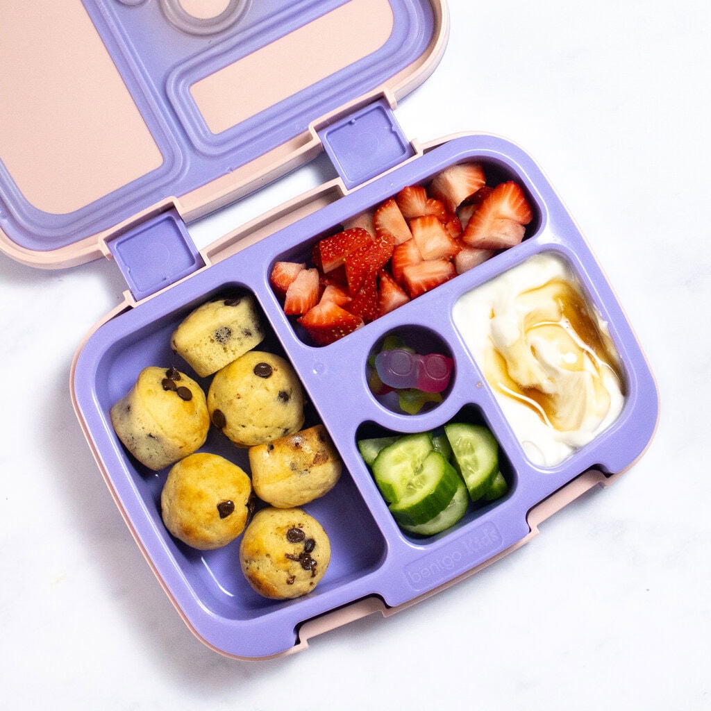 A pink and purple lunchbox sitting on my counter with pancake mini bites, sliced strawberries, cucumbers, gummies and a yogurt and maple syrup dip.