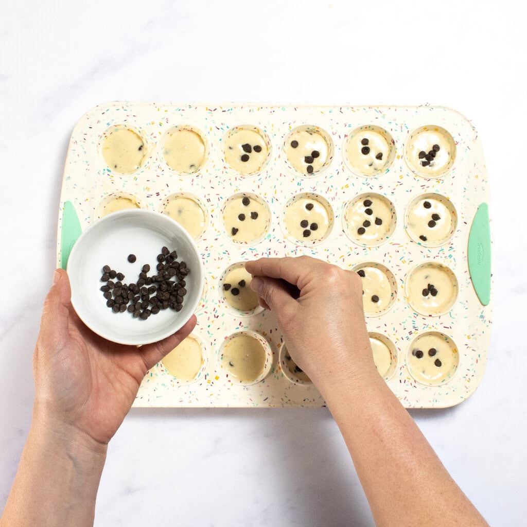 A mini muffin pan with mini pancake bites ready to go, with my hands reaching over to add in a few more chocolate chips on top.