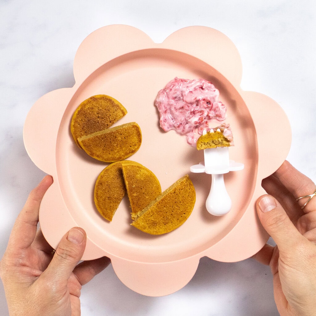 My hands holding a pink baby plate in the shape of a flower, with cut up sweet potato baby pancakes with a dip of raspberries and yogurt.
