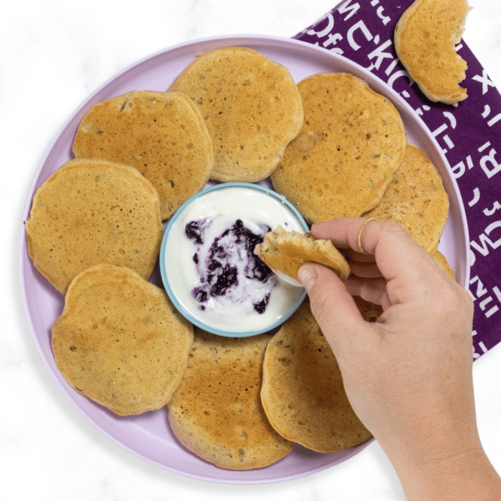 A purple kids plate on a white marble counter with a stack of zucchini pancakes fanned out with a hand holding one that is bitten into over the plate.