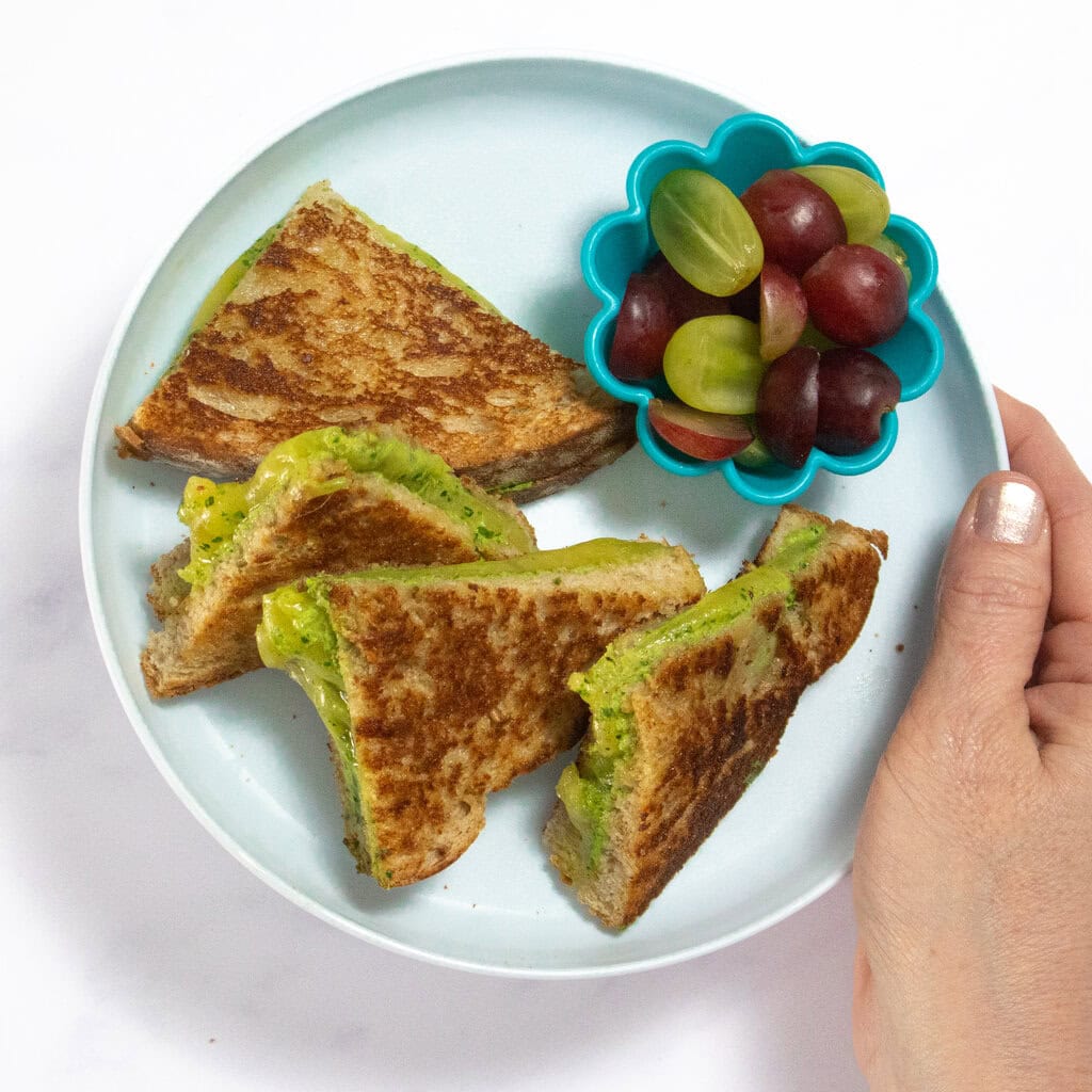 I'm holding a light blue kids plate over my white kitchen counter. On the plate is quartered grilled cheese and a small blue bowl of cut grapes. 